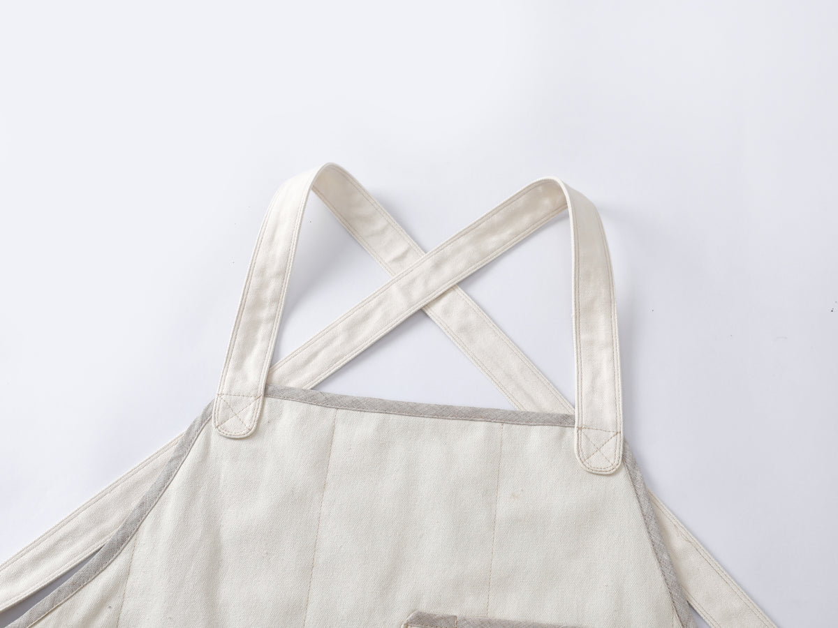 Close-up of a light beige canvas chef apron with wide adjustable shoulder straps crossing at the neck, shown against a plain white background.