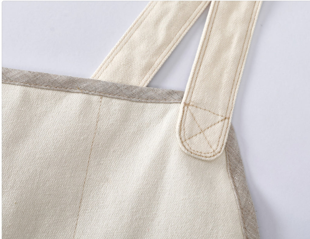 Close-up detail of a canvas chef apron showing the off-white woven fabric body, a flax linen fabric trim along the edge, and a shoulder strap with strong hand-stitching.