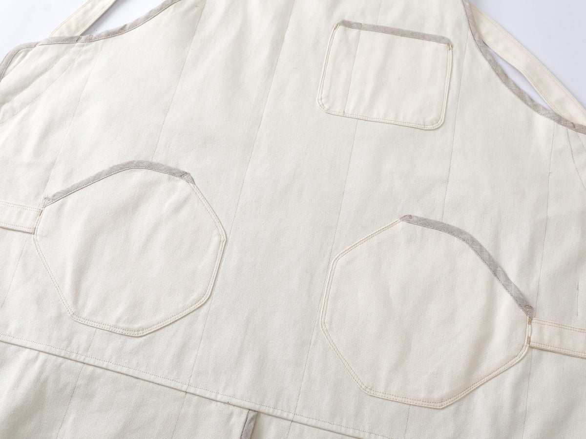 A close-up of a cream-colored canvas chef-apron with octagonal and rectangular pockets, showing the texture of the fabric and the contrast stitching.
