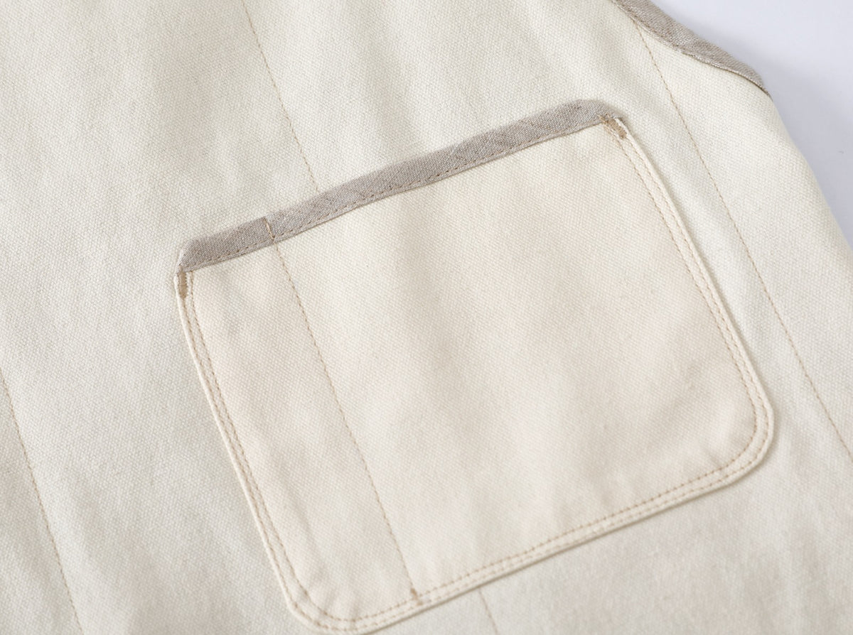 Close-up detail of a cream-colored canvas-linen chef-apron featuring a large patch chest pocket with contrasting pure flax linen trim.