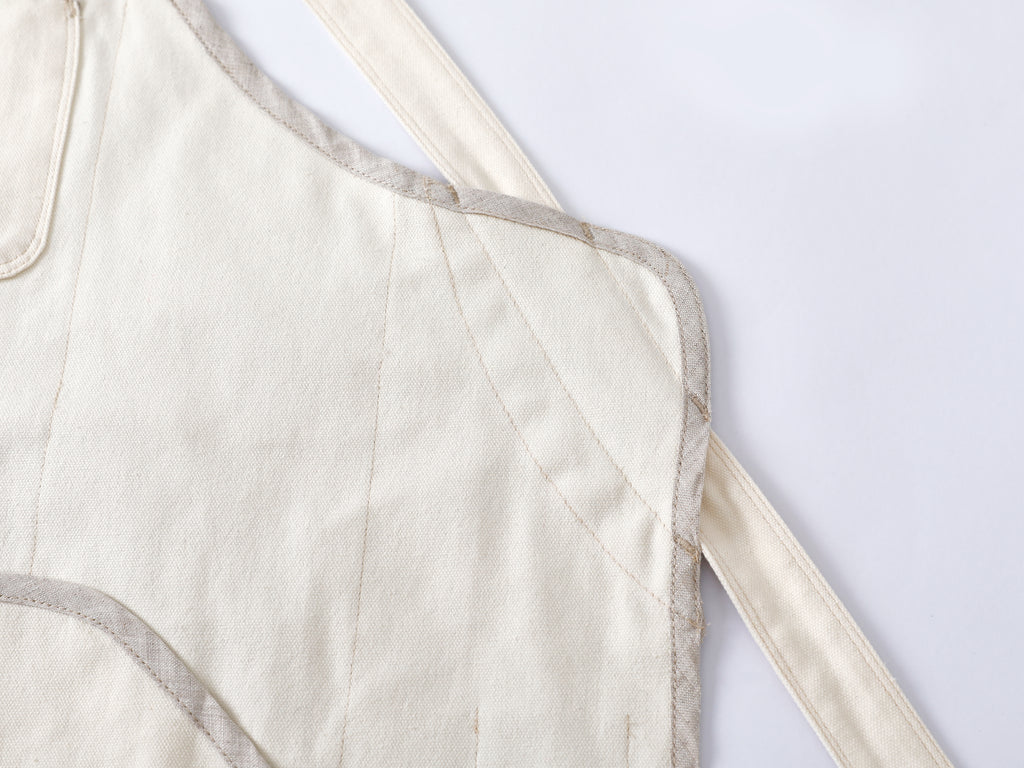 Close-up of a cream-colored quilted fabric chef apron with pure flax linen trim and cotton canvas straps on a white background.