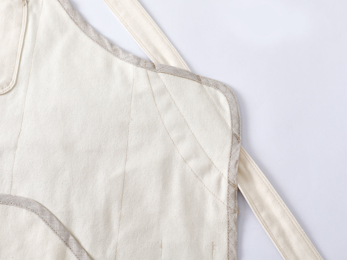 Close-up of a cream-colored quilted fabric chef apron with pure flax linen trim and cotton canvas straps on a white background.